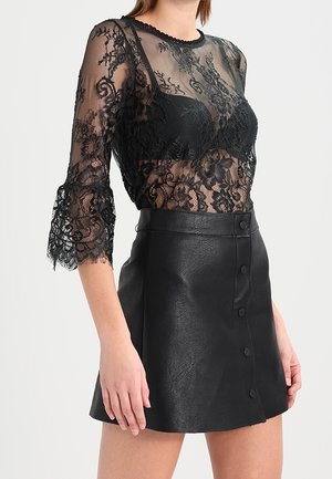 Frau trägt ein schwarzes, transparentes Spitzentop mit Blumenmuster und einen schwarzen, knopfbesetzten Minirock aus Leder.