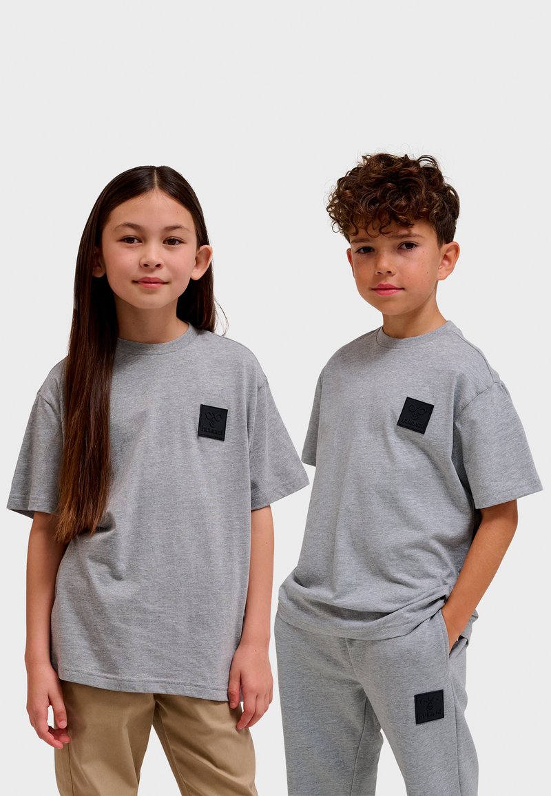 Två barn bär matchande oversized grå t-shirts i mjuk bomull, med en fyrkantig svart logotypmärke på bröstet. Pojkens outfit inkluderar grå sweatpants.