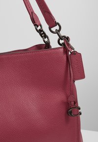 Gros plan d’un sac à main en cuir rose foncé texturé avec double bandoulière et attaches en anneaux de chaîne métallique, avec une étiquette et une petite fermeture métallique.