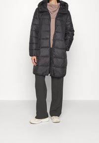 Černý prošívaný kabát s kapucí, s prošitým vzorem, předním zipem a dlouhým střihem, nošený přes béžový top a šedé kalhoty.