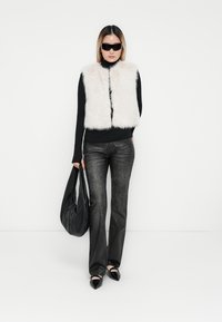 Gilet en fausse fourrure couleur crème pâle, col roulé noir, jeans noirs et ballerines noires à bouts pointus. Portant un grand sac noir décontracté.