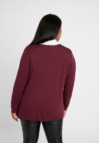 Femme aux longs cheveux noirs portant un haut à manches longues bordeaux, avec un col blanc visible, et un pantalon en cuir noir, se tenant devant un fond uni.