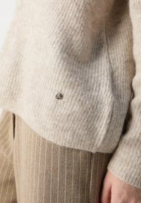Beige knit sweater with a ribbed texture, featuring a small silver emblem on the hem, paired with brown striped trousers.
