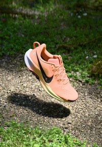 Baskets de course Nike en pêche doux, tige en mesh léger, grand swoosh noir, semelle texturée et talon rehaussé. Flottant sur un fond naturel.