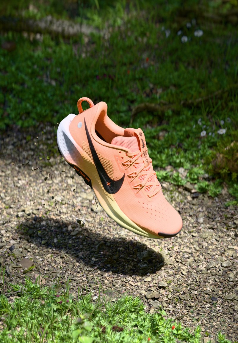 Zapatilla de running de Nike en melocotón suave, parte superior de malla ligera, prominente swoosh negro, suela texturizada y talón elevado. Flotando contra un fondo natural.