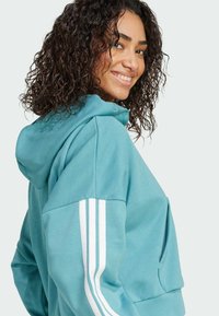 Teal huppari, jossa on iso huppu ja valkoiset kolme raitaa hihansuissa. Pehmeä rakenne, rento leikkaus ja etutasku.