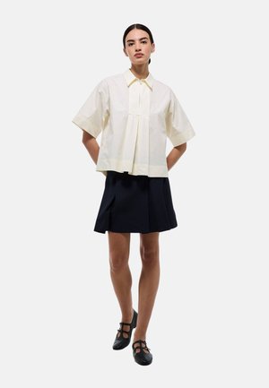 Chemise blanche à col, à manches courtes, avec des plis à l'avant, associée à une jupe noire. Chaussures noires avec des détails de bride.