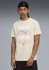 Lys beige bomulds t-shirt med et flerfarvet Puma logo design, korte ærmer og en løs pasform. Klassisk crew-udskæring.