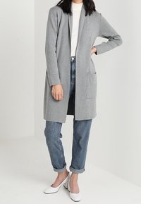 Grauer langer Cardigan mit offener Vorderseite, zwei vorderen Taschen, getragen über einem weißen strukturierten Oberteil und blauen Jeans mit umgeschlagenem Saum, kombiniert mit weißen Absätzen.