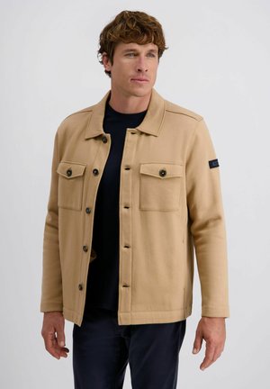 Beige Jacke aus weichem Stoff mit Kragen, zwei Fronttaschen und schwarzen Akzenten an den Ärmeln. Die Knöpfe sind dunkel.