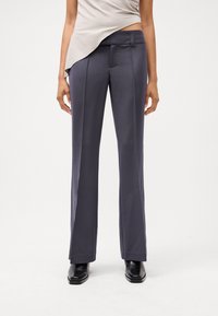 Pantaloni larghi grigio scuro con una texture liscia, dotati di pieghe frontali e una vita piatta. Abbinati a un top di colore chiaro.