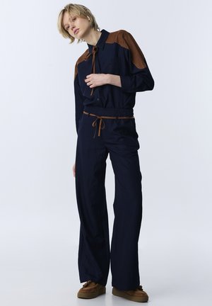 Giovane con capelli biondi corti che indossa una tuta blu navy con toppe marroni sulle spalle e scarpe marroni, in piedi davanti a uno sfondo semplice.