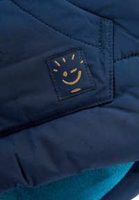 Veste matelassée bleu marine avec une couche extérieure texturée, présentant un logo de visage souriant en or sur un patch en tissu, et une doublure turquoise douce.