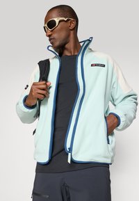 Hombre que lleva una chaqueta de forro polar azul claro y blanca con ribete azul, camisa negra, pantalones oscuros, gafas de sol deportivas beige y lleva una mochila negra.