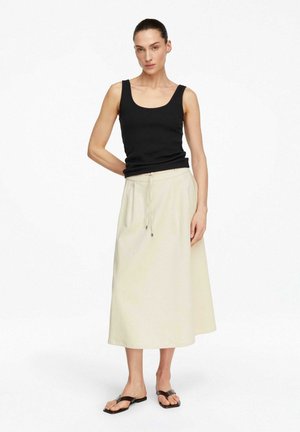 Kobieta o ciemnych włosach w czarnym topie bez rękawów, beżowej spódnicy midi z troczkiem i czarnych sandałach, stojąca na prostym białym tle.