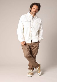 Chemise blanche en velours côtelé avec deux poches avant, associée à un pantalon cargo marron et des baskets beiges avec des semelles blanches. Tenue décontractée.