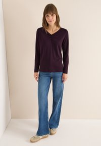 Long-sleeve, deep purple top with a V-neck and gathered detail. Paired with light blue wide-leg jeans and light-colored sneakers.