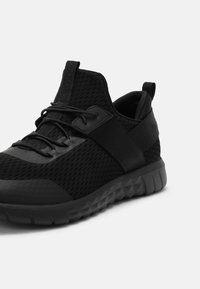 Černá sportovní sneaker s prodyšným svrškem, elastickým páskem přes střední část nohy, texturovanou podrážkou a poutkem na jazyku a patě.