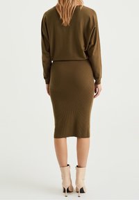 Robe en tricot côtelé vert olive avec des manches longues, une silhouette ajustée et une longueur au genou. Portée avec des bottines à talons beiges.