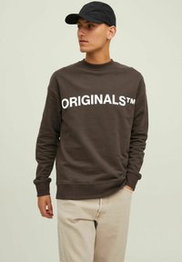 Brown sweatshirt with "ORIGINALS™" in large white letters, round neckline, and ribbed cuffs, paired with light beige pants.