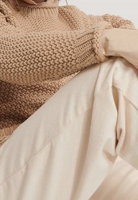 Beige gestrickter Pullover mit hohem Kragen und gerippten Bündchen, kombiniert mit off-white, lässigen Hose in lockerer Passform. Weiche Textur und lässiger Look.