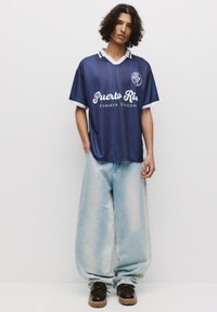 T-shirt rayée marine avec col et poignets blancs, arborant "Puerto Ris" et "SAISON D'ÉTÉ" en lettres blanches en gras. Jeans en denim bleu clair, coupe ample.