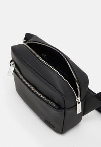 Borsa a spalla in tessuto nero con superficie testurizzata, chiusure a doppia zip e tracolla regolabile. Presenta una tasca frontale con dettagli in metallo.