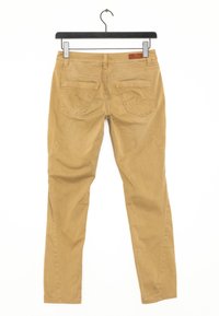 Pantalon ajusté de couleur sable avec une texture lisse, doté d'un design à cinq poches et de poches arrière aux coutures inclinées, suspendu à un cintre noir.