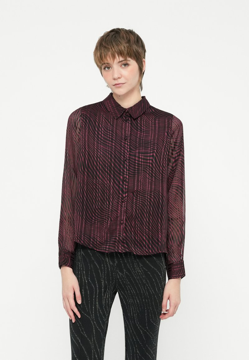 Blusa trasparente di un rosso scuro con un sottile motivo a onde, maniche lunghe, chiusura frontale con bottoni e colletto classico. Abbinata a pantaloni neri con texture.