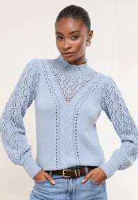 Heled sinine kootud kampsun pitsiliste mustritega, kõrge krae ja ribakäistega. Kanda tume vöö ja sinised denim püksid.
