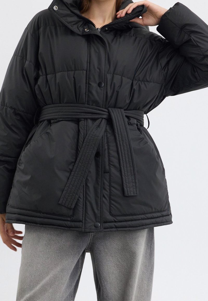 Femme portant une veste matelassée noire avec une ceinture nouée à la taille et un col montant, associée à un pantalon gris clair.