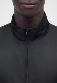Gros plan d'une personne portant une veste noire zippée jusqu'au col, ne montrant que la partie inférieure du visage et le cou.
