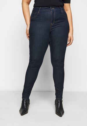Femme portant un jean skinny bleu foncé et des bottines noires à bout pointu, debout devant un fond uni.