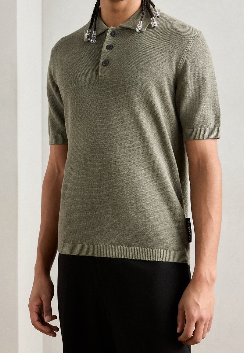 Homme portant un polo tricoté kaki à manches courtes avec trois boutons et un pantalon noir, debout sur un fond neutre.