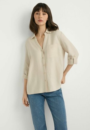 Kobieta o włosach do ramion, ubrana w beżową koszulę na guziki z kieszonką i podwiniętymi rękawami oraz niebieskie dżinsy, stojąca przy białej ścianie.