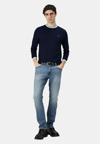 Giovane uomo che indossa un maglione blu navy con rifiniture bianche, jeans azzurri chiari, cintura nera e scarpe nere, in piedi con le mani nelle tasche su uno sfondo bianco.