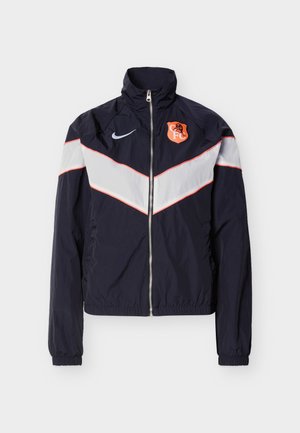 Temno modra športna jakna na zadrgo z belimi črtami v obliki čevrona, elastičnimi manšetami, logotipom Nike na desnem prsnem delu in oranžnim grbom ekipe na levem prsnem delu.