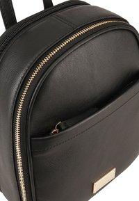 Sac à dos en cuir noir avec une forme arrondie, des détails de fermeture éclair dorés, une poche zippée à l'avant et un petit accent de logo métallique.