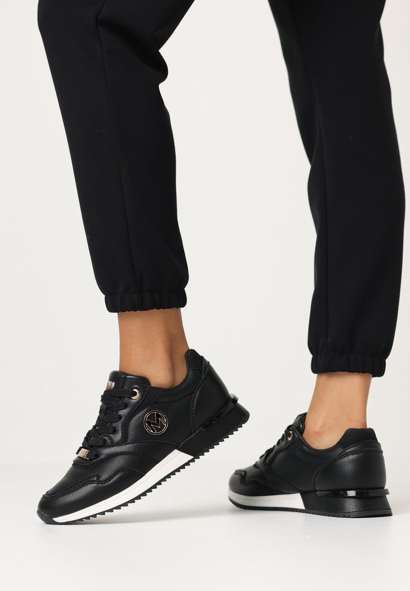 Personne portant des baskets en cuir noires avec des semelles blanches et un pantalon de jogging noir, debout sur une surface blanche.