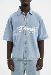Lyseblå denim skjorte med korte ærmer, hvidt logo, knaplukning foran og to sidelommer. Har en afslappet pasform og let tekstureret stof.