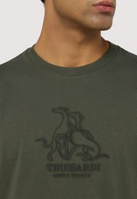 Olivově zelené bavlněné tričko s kulatým výstřihem, zdobené černou vyšitou grafikou tří psů a textem "TRUSSARDI GENTLE SOCIETY" níže.
