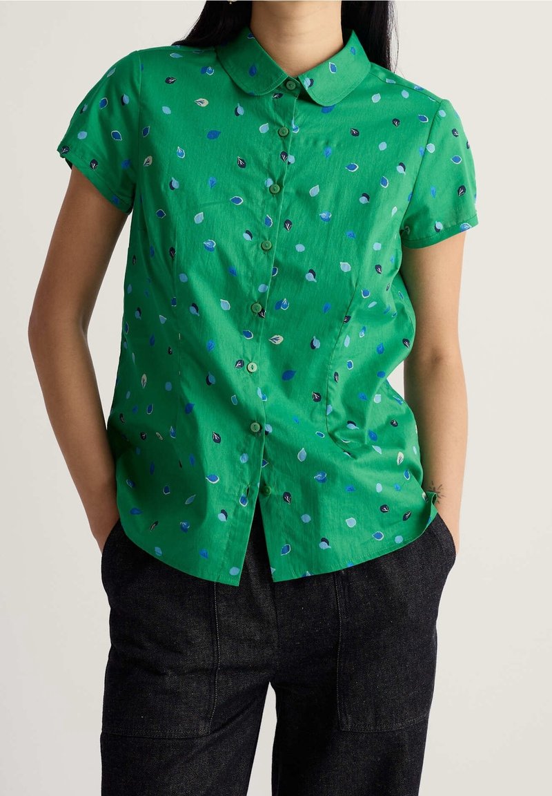 Femme portant une chemise verte à manches courtes avec des motifs de feuilles bleues et blanches et un pantalon foncé, les mains dans les poches.