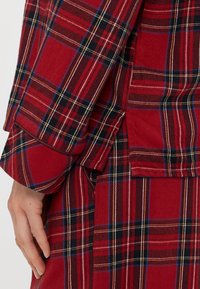 Czerwony materiał w kratę z teksturowanej tkaniny z czarnymi, niebieskimi i żółtymi paskami. Posiada długie rękawy i boczny rozcięcie.