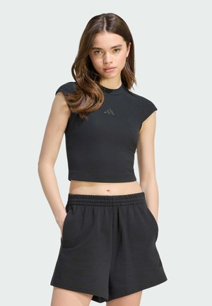 Ung kvinde med langt brunt hår iført sort ærmeløst croptop og sorte kort elastiske shorts i taljen, hænder i lommerne, neutralt baggrund.