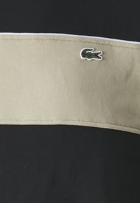 Maglietta nera con una ampia striscia orizzontale beige, caratterizzata da un piccolo logo di un coccodrillo ricamato in verde e bianco centrato sulla striscia.