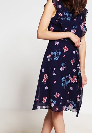 Kukkakuvioinen mekko, jossa tummansininen tausta, kevyt kangas, röyhelöyksityiskohdat hartioilla ja polven pituinen A-linjainen siluetti.