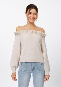 Light beige off-the-shoulder sweater with ruffle detailing, long sleeves, and ribbed hem. Soft texture and relaxed fit. Worn with light blue jeans.