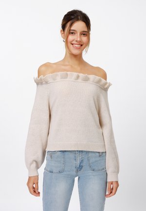 Pull léger beige clair à épaules dénudées avec des volants, manches longues et ourlet côtelé. Texture douce et coupe décontractée. Porté avec un jean bleu clair.