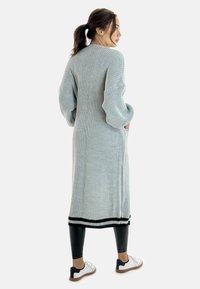 Lungo cardigan lavorato a maglia grigio chiaro con tessuto testurizzato, maniche a coste, tasche laterali e bordi neri, indossato sopra leggings neri e scarpe bianche.