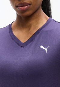 Κοντινό πλάνο ενός ατόμου που φοράει μοβ αθλητική μπλούζα με V λαιμόκοψη και λευκό λογότυπο Puma στο στήθος.